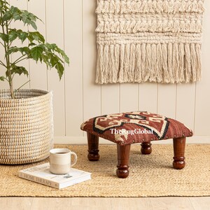 May include: A square footstool with a woven fabric top featuring a geometric pattern in rust, cream, and black, supported by four dark brown turned wooden legs. The fabric is secured with metal studs. The text TheRugGlobal is printed on the top.