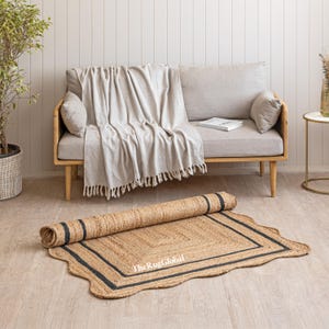 May include: A rectangular natural jute rug with scalloped edges and two black border lines. The rug is partially rolled up, revealing a woven texture. The text TheRugGlobal is printed in white on the surface of the tan and black fiber rug.