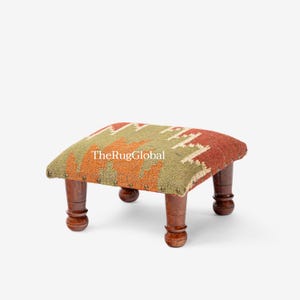 Può includere: Un poggiapiedi basso con piano rettangolare rivestito in tessuto intrecciato con motivo geometrico nei toni del verde oliva e dell'arancione bruciato. Il testo TheRugGlobal è stampato sopra. Ha quattro gambe in legno tornito marrone scuro.