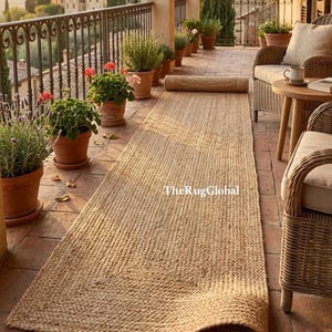 Puede incluir: Una alfombra pasillera rectangular de yute en color natural con textura trenzada, que presenta el texto TheRugGlobal impreso en su superficie. La alfombra está colocada sobre un balcón de baldosas de terracota, entre plantas y muebles de mimbre.