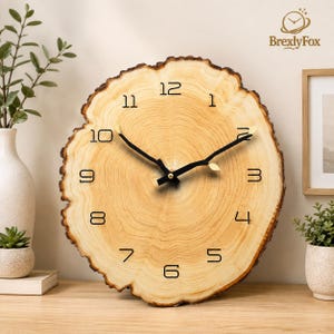 May include: A round wall clock made from a natural wood slice with visible tree rings and bark edges. The clock face features black Arabic numerals and black metal hands with small gold leaf accents. The brand name BrexlyFox is printed in the top right corner.