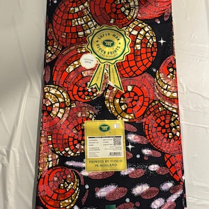 Può includere: Un pezzo di tessuto in raso di cotone piegato con sfondo nero e un motivo ripetitivo di cerchi a mosaico rossi, arancioni e dorati. Un'etichetta gialla riporta "Printed by Vlisco in Holland" e un sigillo verde reca la scritta "Super-Wax".