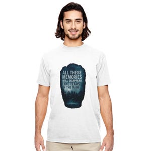 May include: White t-shirt featuring a dark blue graphic with the text "ALL THESE MEMORIES WILL DISAPPEAR LIKE TEARS IN THE RAIN". The graphic depicts a cityscape silhouette. The shirt is worn by a person.