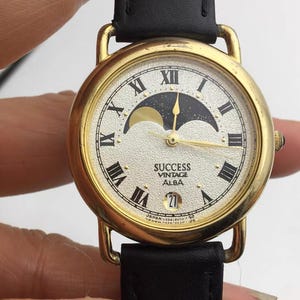 May include: Vintage Alba Success watch with a gold-tone case and a black leather strap. The watch face displays Roman numerals, a moon phase design, and the date. The watch is marked "Japan" and "Vintage Alba Success".