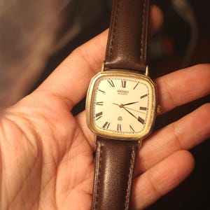 May include: A gold-tone Seiko Quartz wristwatch featuring a square-shaped case with rounded corners, a cream-colored dial with black Roman numeral hour markers, and a dark brown leather strap with visible stitching along the edges.