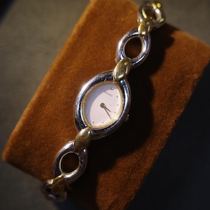 May include: A Seiko wristwatch featuring an oval-shaped white dial with gold-tone hands and markers. The watch band is constructed from alternating silver-tone and gold-tone oval links, creating a continuous chain design that secures the timepiece.