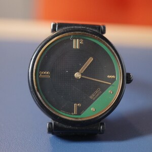 May include: A black Seiko Quartz watch with a round face and a black strap. The watch face has a black background with gold hands and markers, and a green border. The watch is set against a blue background.