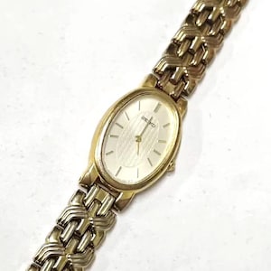 Può includere: Orologio da polso Seiko color oro con quadrante ovale e bracciale a maglie abbinato. Il quadrante dell'orologio è bianco con indici e lancette neri. Il bracciale presenta un motivo ripetuto di maglie.