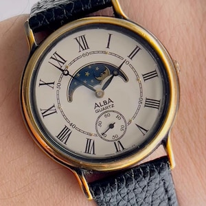 May include: An Alba Quartz wristwatch with a gold-tone round case and a textured black leather strap. The off-white dial features black Roman numeral hour markers, a moon phase sub-dial at the top, and a small seconds sub-dial at the bottom.