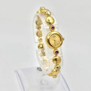 Puede incluir: Un reloj de pulsera Seiko dorado con esfera redonda y pulsera de cuentas. La esfera del reloj es dorada con agujas y marcadores negros. La pulsera presenta cuentas doradas y plateadas, con detalles en rojo.
