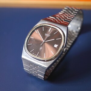 Op de afbeelding: Een vintage Seiko Quartz horloge met een zilverkleurige roestvrijstalen band en kast. De wijzerplaat van het horloge is bruin met zilveren wijzers en markeringen. Het horloge heeft een datumweergave op 3 uur. Het horloge ligt op een blauw oppervlak.
