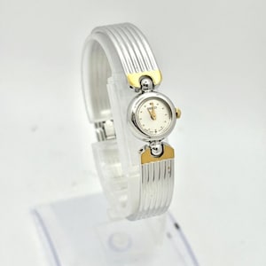 May include: A silver-toned wristwatch with a white face and gold accents. The watch features a round face with gold hands and markers. The band is a silver ribbed design. The watch is displayed on a clear stand.