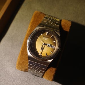 May include: A silver-tone Seiko Quartz Type II wristwatch with a metallic gold sunburst dial, silver hands, and a day-date window. The watch features a textured silver metal link bracelet and a round case with a polished finish.