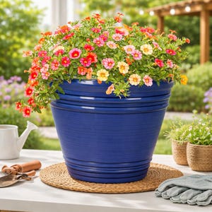 May include: A large, cobalt blue ceramic flower pot filled with colorful flowers in shades of red, pink, and yellow. The pot has a textured surface and sits on a woven mat. Gardening tools and gloves are nearby, suggesting a garden theme.