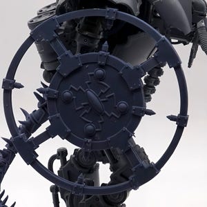May include: A dark gray, mechanical model with a circular shield and spiky details. The shield has an intricate design in the center and is connected to a wheel-like structure. The model appears to be part of a larger, complex structure.