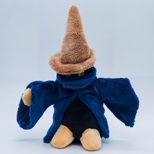 Puede incluir: Un peluche de mago con un sombrero marrón en forma de cono y una túnica azul oscuro. El juguete tiene manos y pies de color beige y un cuerpo negro. El juguete del mago está de pie sobre un fondo blanco.