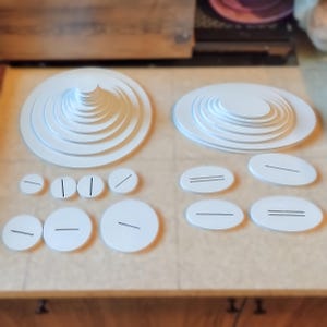 May include: White, layered, circular objects of varying sizes are arranged on a light brown surface. Two larger, tiered, circular structures are present, along with smaller, flat, circular and oval pieces. Some pieces have a single or double line etched across them.