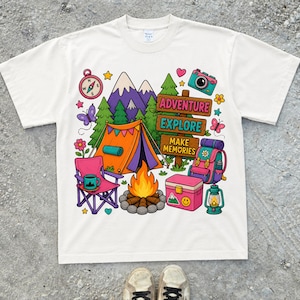 May include: White t-shirt featuring a camping-themed illustration. The design includes a tent, mountains, a compass, a camera, and the words "Adventure, Explore, Make Memories." Other elements include a campfire, backpack, and a lantern.