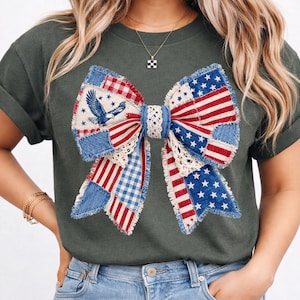 May include: Dark green t-shirt with a patriotic bow design. The bow is made of patchwork fabric with red, white, and blue patterns, including stars, stripes, and an eagle. The bow is centered on the shirt.