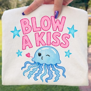 May include: A white, square fabric item with the words "BLOW A KISS" in pink bubble letters, along with a winking blue jellyfish illustration and blue star accents. A small pink heart is also present.