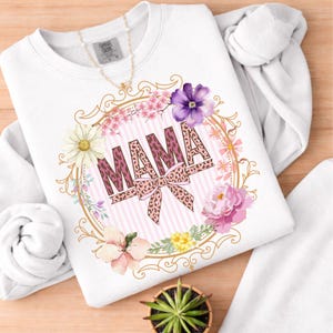 May include: White sweatshirt featuring the word "MAMA" in a leopard print design, encircled by a floral wreath with pink and purple blooms. A gold necklace is draped over the garment.