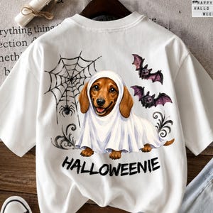 Puede incluir: Una camiseta blanca con el gráfico de un perro salchicha marrón vestido con un disfraz de fantasma blanco. El diseño incluye una telaraña negra con una araña, dos murciélagos morados y negros, y el texto negro HALLOWEENIE bajo el perro.