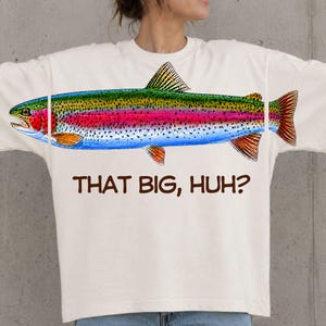 Peut inclure: Un t-shirt à manches courtes blanc cassé présentant une grande illustration colorée d'une truite arc-en-ciel sur la poitrine. Sous le poisson, le texte "THAT BIG, HUH?" est imprimé en police sans empattement marron foncé sur le devant.