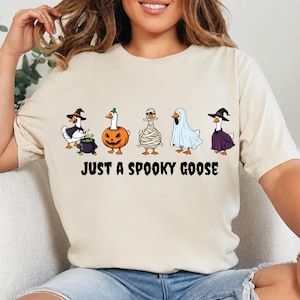 May include: A cream-colored t-shirt featuring a graphic of five geese in various Halloween costumes, including a witch, a pumpkin, a mummy, a ghost, and a wizard. Below the geese, the text reads JUST A SPOOKY GOOSE in black capital letters.