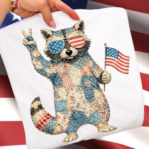 Pode incluir: Uma almofada decorativa quadrada branca com um guaxinim usando óculos de sol com o design da bandeira americana. O guaxinim está segurando uma pequena bandeira americana e fazendo o sinal de paz. O corpo do guaxinim é feito de tecido de retalhos.