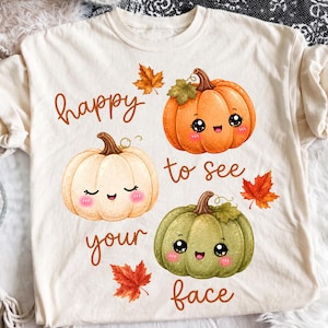 以下が含まれることがあります： クリーム色のTシャツに、オレンジ、白、緑の笑顔の漫画風カボチャが3つ描かれています。茶色の筆記体で「happy to see your face」という文字がプリントされ、オレンジや茶色の秋の紅葉が散りばめられています。