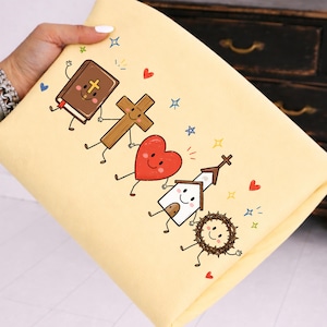 Op de afbeelding: Een lichtgele stof met een cartoonafbeelding van een bruin boek met een gouden kruis, een houten kruis, een rood hart, een wit kerkgebouw en een bruine doornenkroon, allemaal met lachende gezichten, omringd door kleine kleurrijke hartjes en sterren.