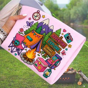 May include: A pink camping-themed design with a tent, campfire, and the words "Adventure, Explore, Make Memories." Other elements include a compass, camera, and backpack. The design is on a pink fabric.
