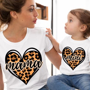 May include: White t-shirts with matching heart designs. One shirt features "mama" in a script font over a leopard print heart, and the other says "mini". The shirts are worn by a mother and child.