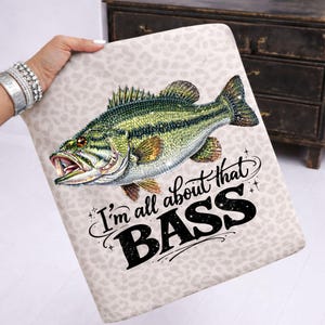 Könnte beinhalten: Ein rechteckiges Kissen mit hellbeigem Leopardenmuster. Eine detaillierte Illustration eines grünen und goldenen Bassfisches ist auf dem Kissen zentriert. Der Text "I'm all about that BASS" steht unter dem Fisch in schwarzer Schrift.