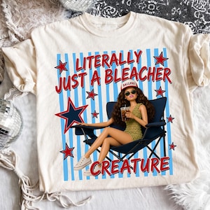 May include: Cream-colored t-shirt with the text "LITERALLY JUST A BLEACHER CREATURE" in red and blue. The graphic includes a doll in a chair, stars, and blue stripes. The doll is wearing sunglasses and a hat.