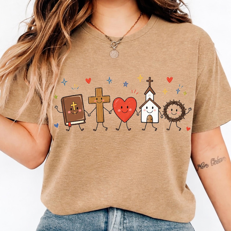 Op de afbeelding: Een beige t-shirt met korte mouwen en een afbeelding van vijf cartoonfiguren die elkaars hand vasthouden: een bruine bijbel, een houten kruis, een rood hart, een wit kerkgebouw en een bruine doornenkroon, versierd met kleine kleurrijke sterren en hartjes.