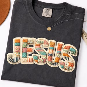 May include: Dark grey t-shirt featuring the word "JESUS" in a patchwork design. The letters are made from various patterned fabrics and edged with cream-coloured lace trim. A small tag is visible near the neckline, and a necklace with a small pendant is draped across the shirt.