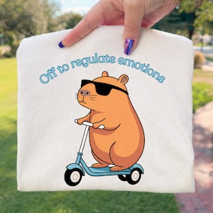 May include: White sweatshirt featuring a cartoon capybara wearing sunglasses and riding a blue scooter. The text "Off to regulate emotions" is arched above the capybara in light blue.