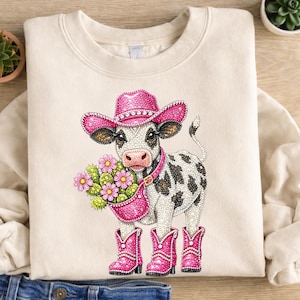 Op de afbeelding: Cr&egrave;mekleurige sweatshirt met een met strass versierde koe met een roze cowboyhoed, laarzen en een halsband. De koe houdt een roze bloemenmand vast. De koe heeft zwart-witte vlekken.
