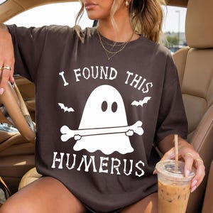 May include: A dark brown short-sleeve t-shirt featuring a white graphic of a ghost holding a bone. The text "I FOUND THIS HUMERUS" is printed in white above and below the ghost, accompanied by two small white bat silhouettes on either side.