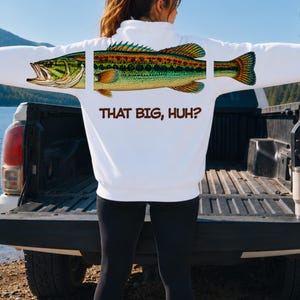Peut inclure: Un sweat-shirt à capuche blanc avec un motif de grand poisson vert et jaune divisé en trois sections sur le dos et les manches. Sous le poisson, le texte "THAT BIG, HUH?" est imprimé en lettres capitales marron, grasses et sans empattement.