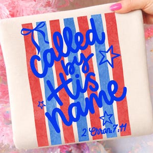 Puede incluir: Un cojín cuadrado color crema con un diseño patriótico. El cojín presenta rayas verticales rojas y azules y las palabras "Called by His Name" en escritura azul, junto con acentos de estrellas y el versículo bíblico 2 Crón. 7:14.
