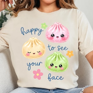 May include: A beige t-shirt featuring a graphic of three cartoon dumplings in pink, yellow, and green, each with a smiling face. The text "happy to see your face" is printed in blue script, accompanied by small pink, yellow, and green flower icons.