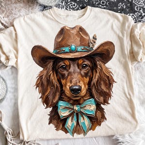 Puede incluir: Una camiseta color crema con el gráfico de un perro salchicha marrón de pelo largo que lleva un sombrero de vaquero marrón con una banda de piedra turquesa y una pluma, y un gran lazo azul claro texturizado alrededor del cuello.