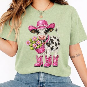 Op de afbeelding: Lichtgroen T-shirt met een koe-illustratie. De koe draagt een roze cowboyhoed, laarzen en een handtas vol bloemen. De koe heeft zwart-witte vlekken. Het ontwerp is gedetailleerd en kleurrijk.