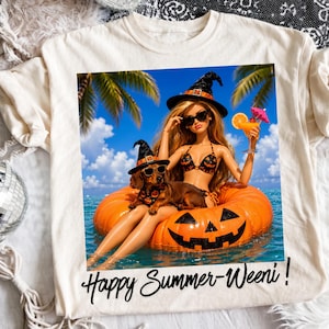 Op de afbeelding: Een crèmekleurig t-shirt met een afbeelding van een pop en een teckel met heksenhoeden en zonnebrillen, drijvend op een pompoen-zwemband in de oceaan. De tekst "Happy Summer-Weeni!" staat in zwarte letters onder de afbeelding gedrukt.