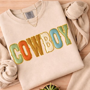 May include: A beige sweatshirt with the word "COWBOY" in colorful, textured lettering. The letters are outlined in gold and filled with shades of orange, blue, green, and cream. A gold necklace with a butterfly pendant is visible near the neckline.