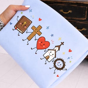 Op de afbeelding: Een lichtblauw sweatshirt met een grafische print van vijf stripfiguren die elkaars hand vasthouden: een bruine Bijbel, een houten kruis, een rood hart, een witte kerk en een bruine doornenkroon, versierd met kleine kleurrijke sterretjes en hartjes.