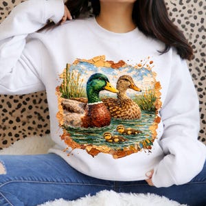 May include: White sweatshirt featuring a detailed painting of a male and female duck with ducklings swimming in water. The ducks have green, brown, and yellow coloring. The artwork is framed with a textured, torn-paper effect.