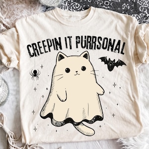Op de afbeelding: Een crèmekleurig T-shirt met korte mouwen en een zwarte afbeelding van een kat verkleed als spook. De tekst "CREEPIN IT PURRSONAL" staat boven de kat, vergezeld van illustraties van een kleine spin, een vleermuis en verschillende sterren.